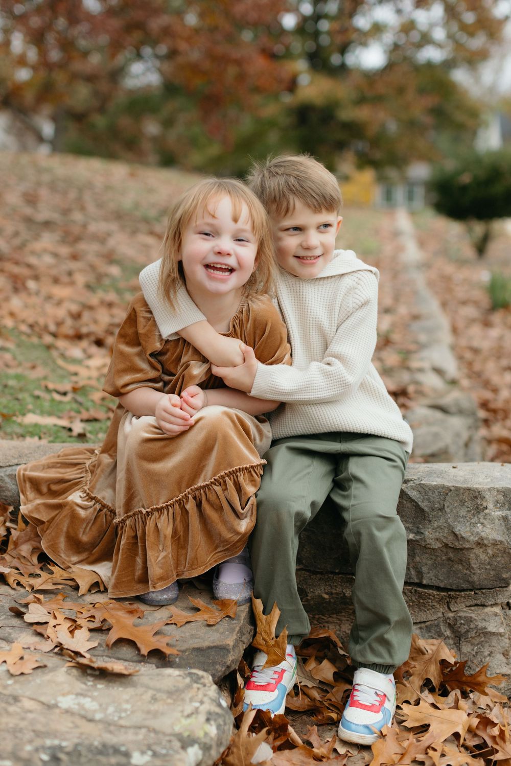 Two children sit together on stone steps surrounded by scattered autumn leaves in matching white and brown outfits.