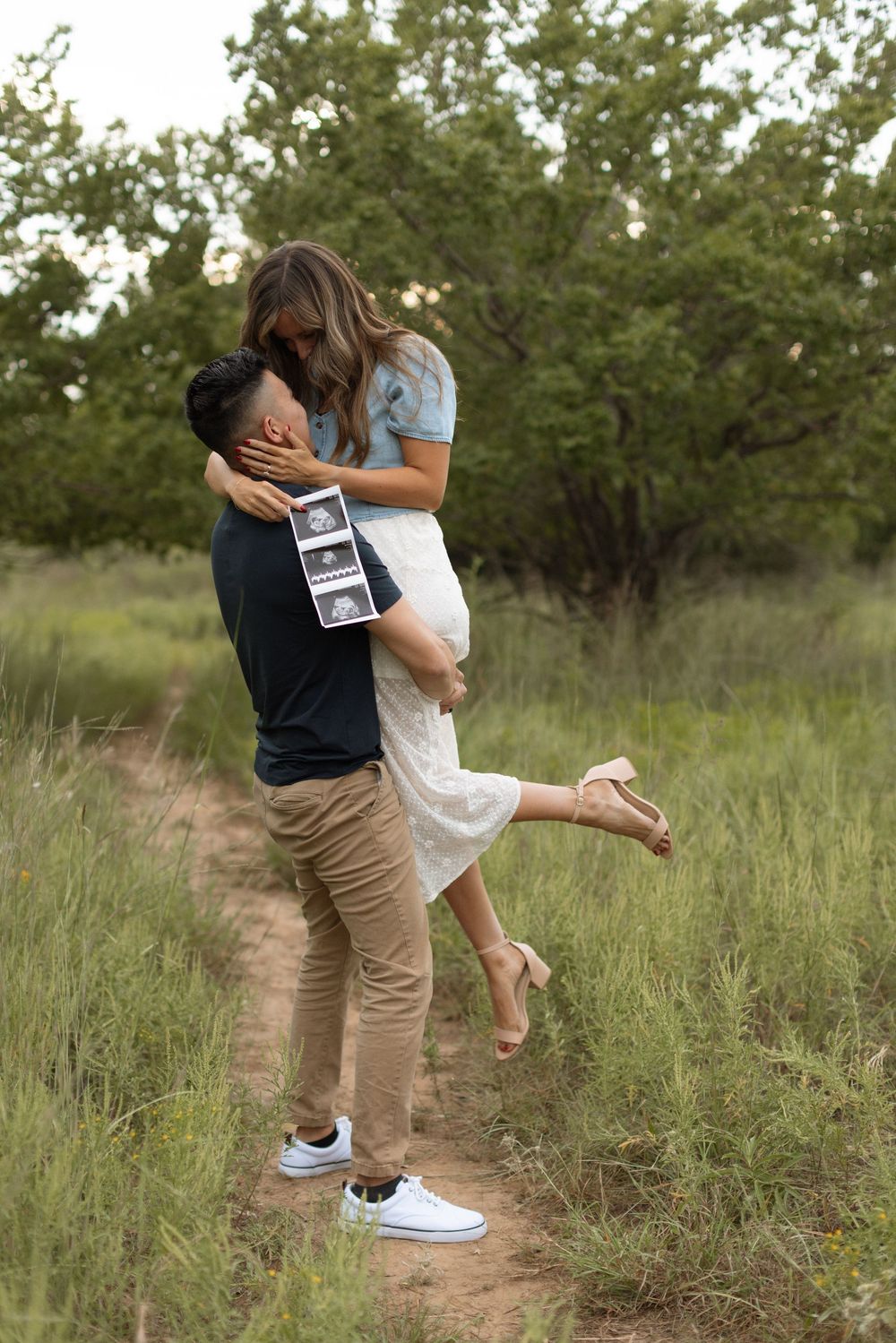 A couple embraces on a dirt path while holding an ultrasound photo in a natural outdoor setting.