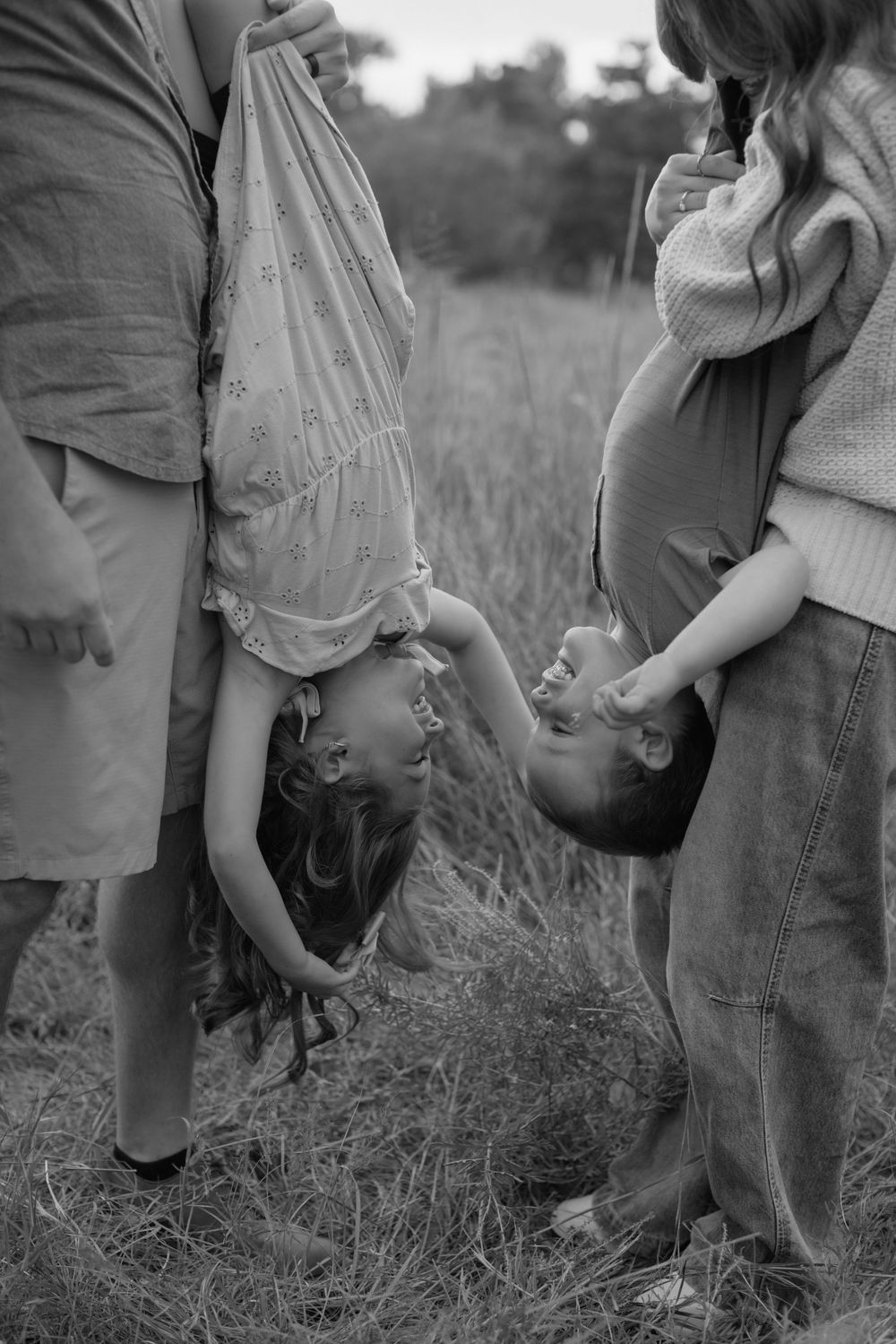 Black and white photo of a pregnant woman in a sweater standing next to a child playfully hanging upside down in a field.