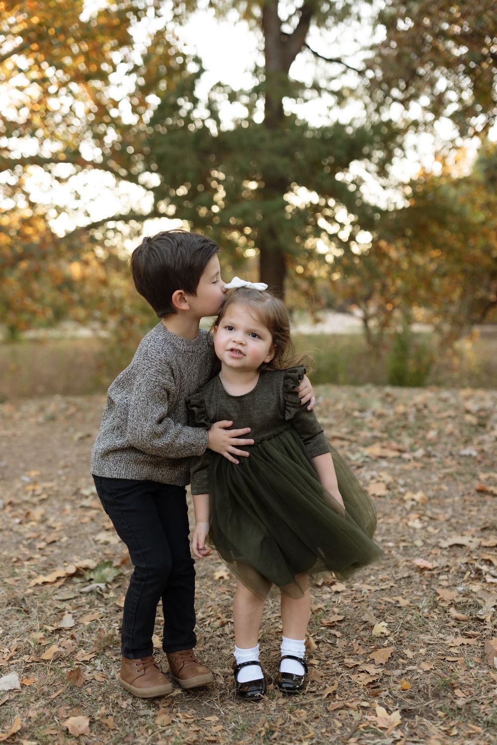 Two children share a sweet moment together in an autumn forest setting with fallen leaves on the ground.