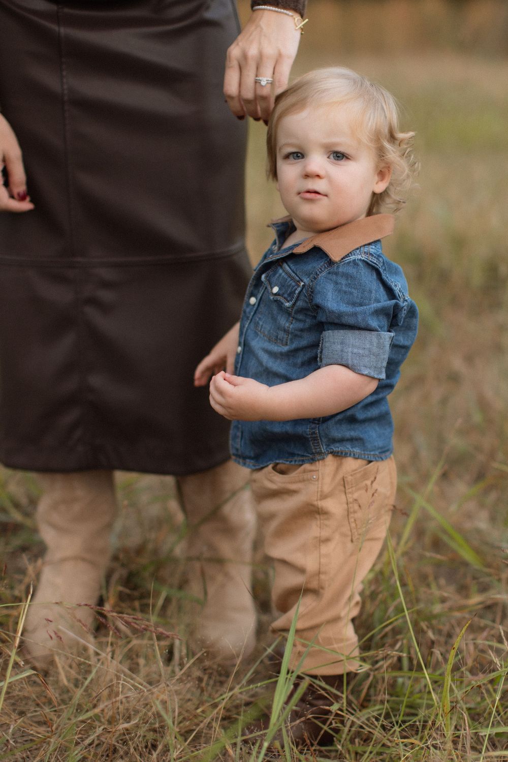 Small child wearing denim overalls and khaki pants stands in grassy outdoor setting with adult legs visible in background.