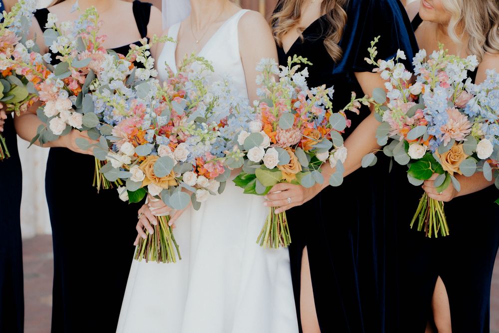 Wedding party stands in a row holding elegant pastel bouquets with blush and cream flowers against a neutral backdrop.