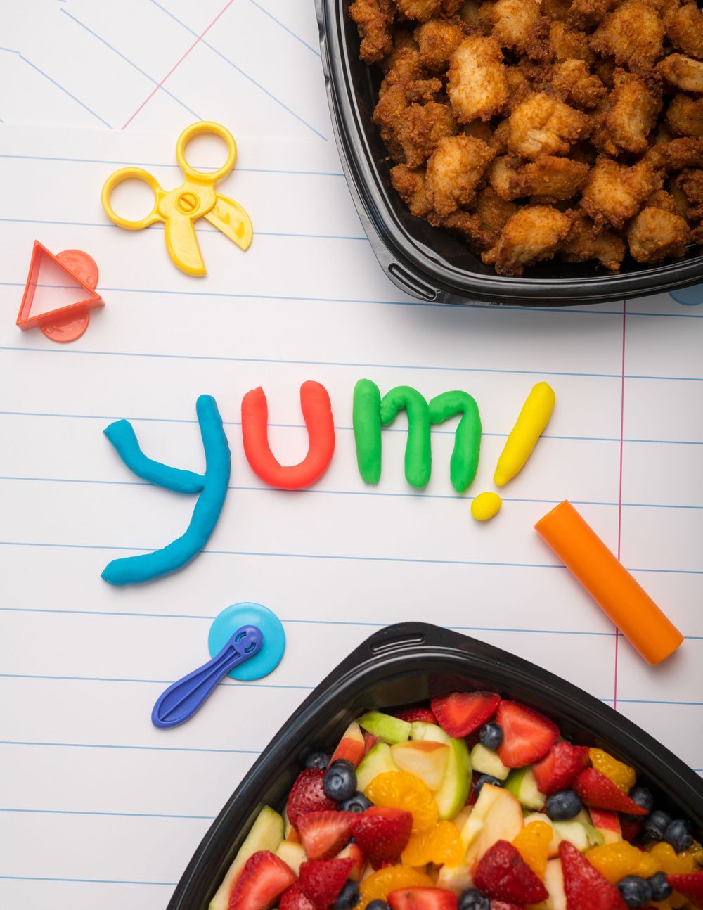 Chicken nuggets and fresh fruit platter with colorful letter magnets spelling 'yum'.