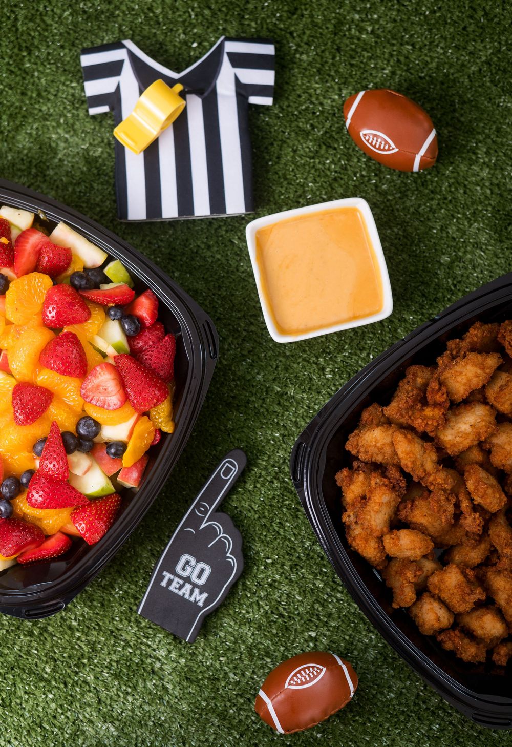Football-themed spread with chicken nuggets, fruit platter, and sauces on artificial turf background.