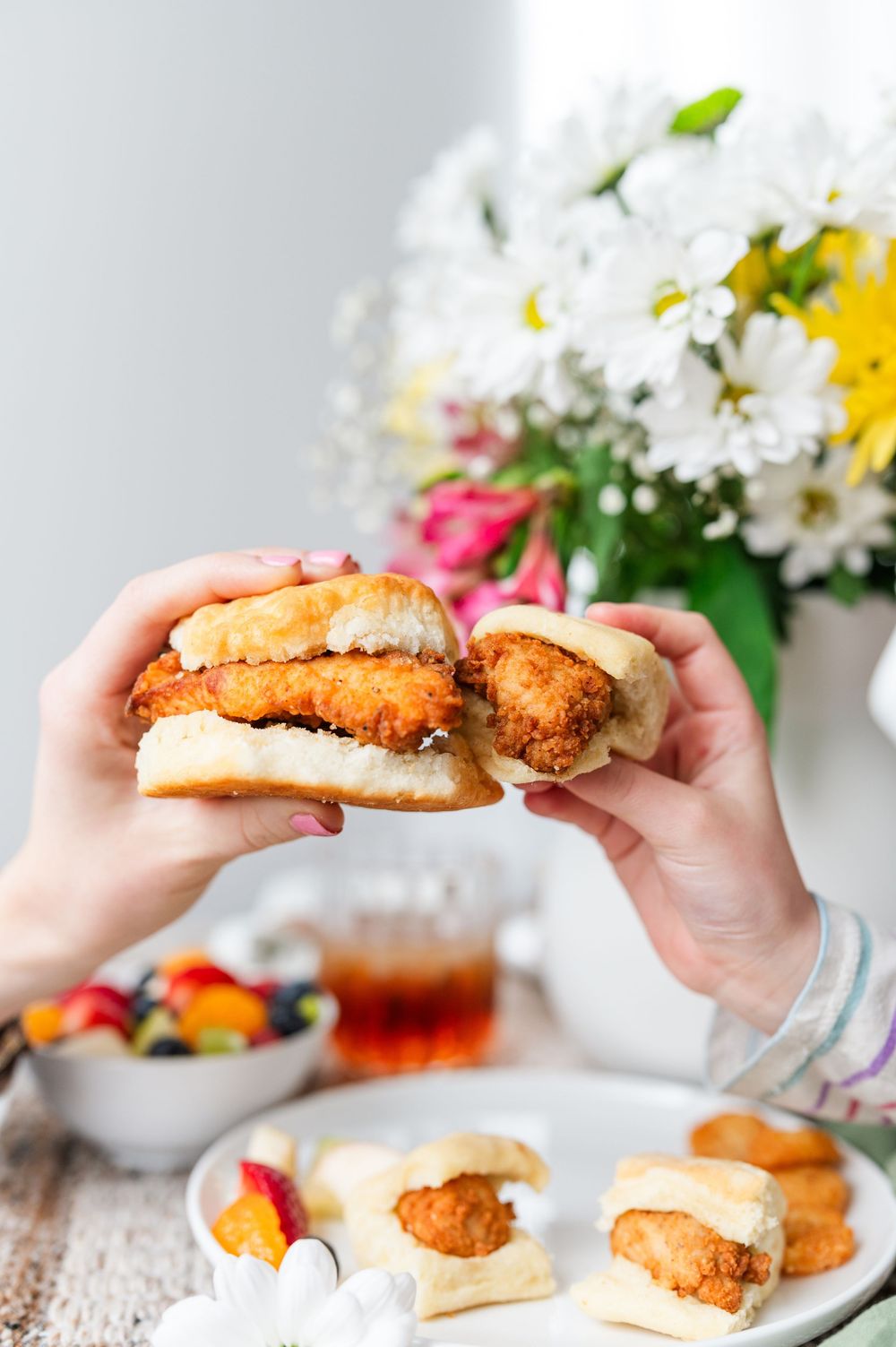Hands holding crispy chicken sandwiches with fresh flowers in background.