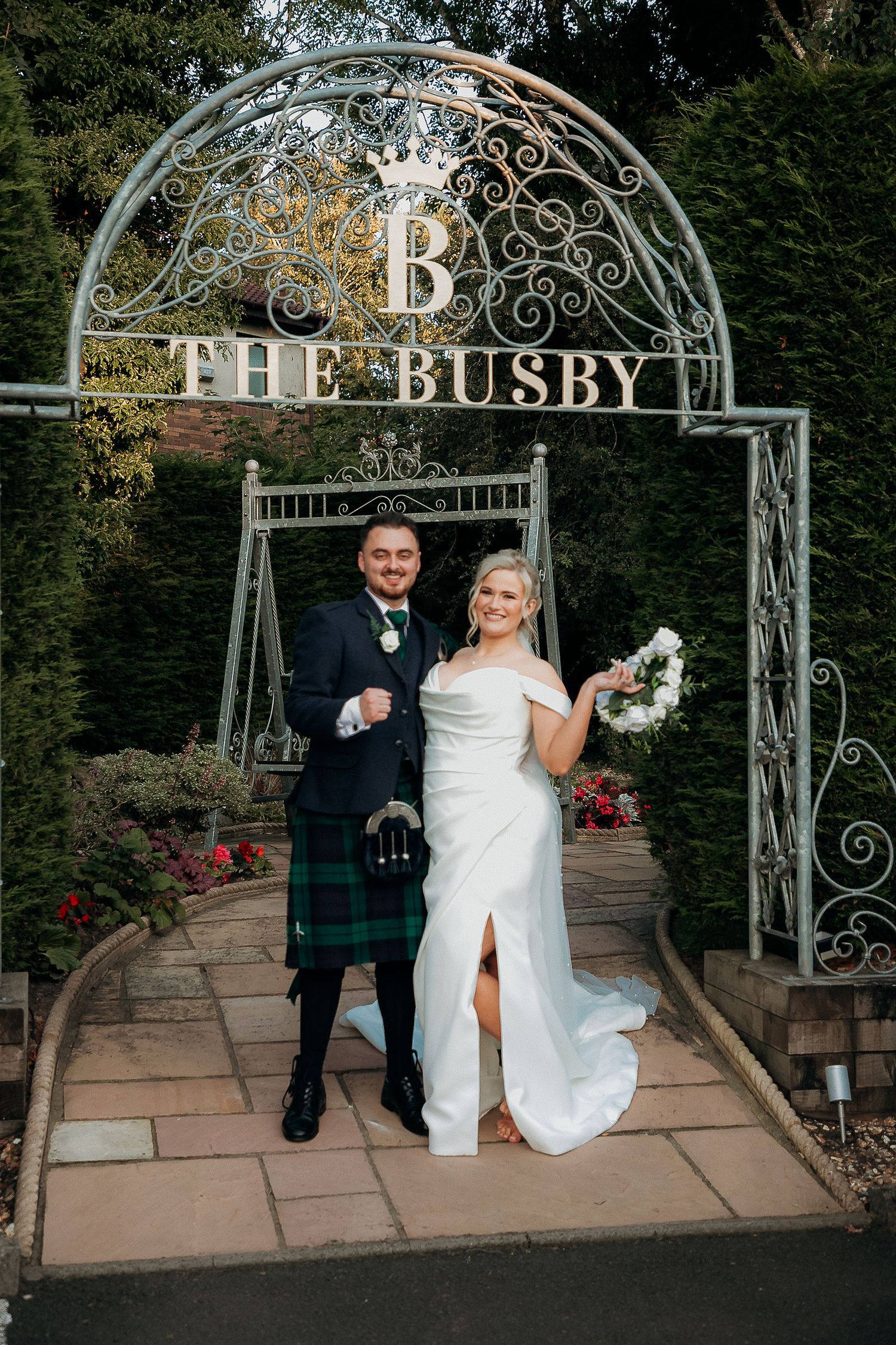 Wedding couple standing under ornate Busby arch entrance in Scottish attire.