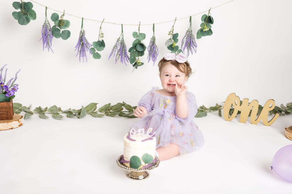 Baby in purple dress sits on white backdrop with eucalyptus garland and wooden birthday cake topper decorations.