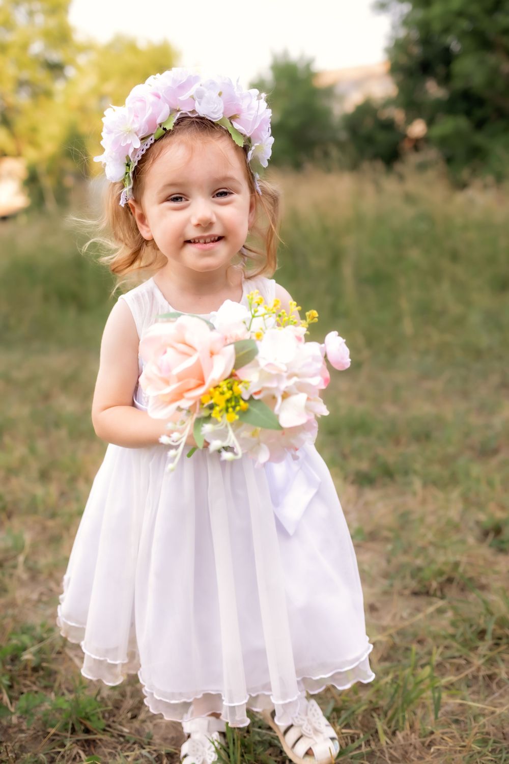 A child in a white dress and floral crown stands in a grassy field holding yellow and pink flowers.