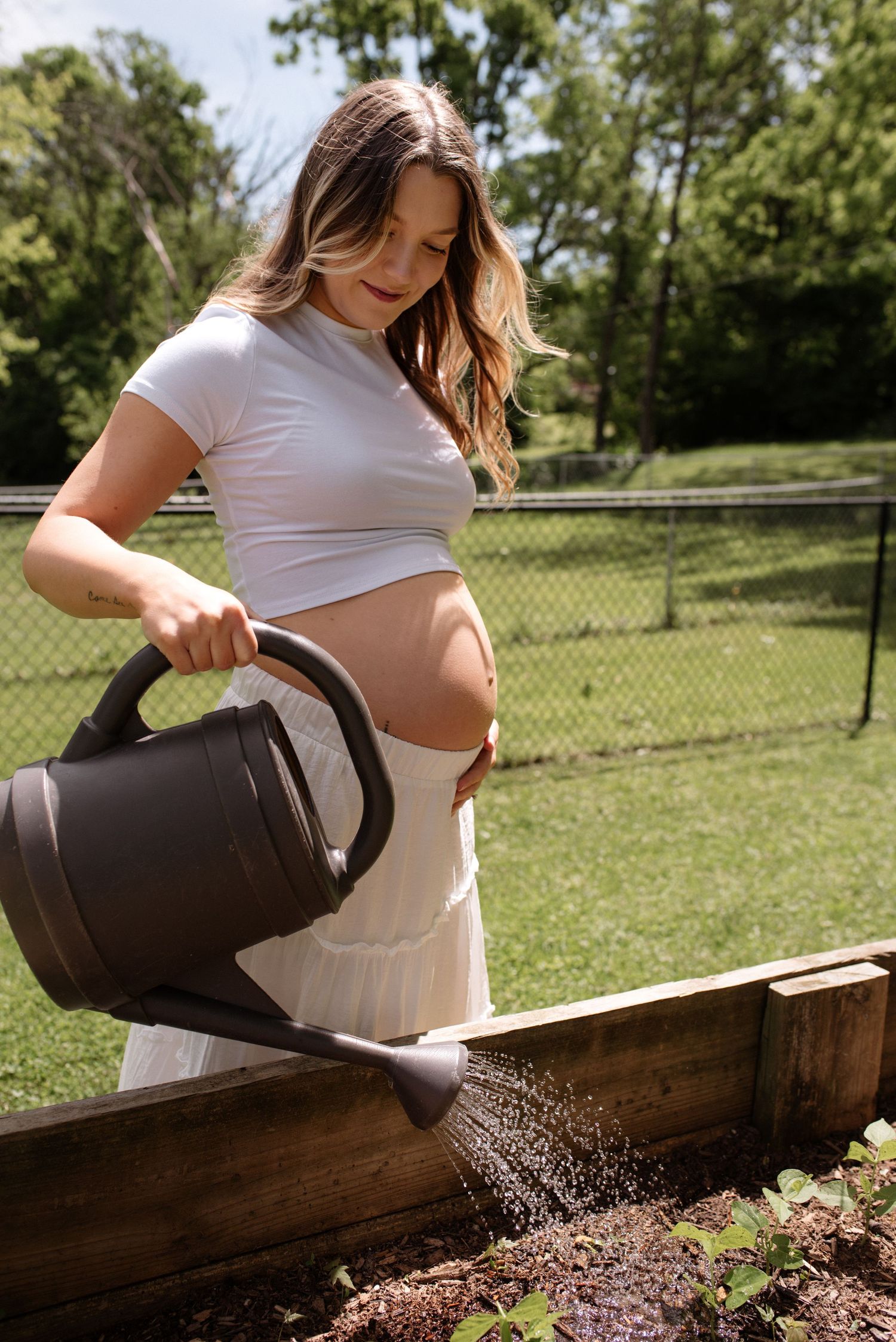 A pregnant woman in a white crop top waters a raised garden bed with a black watering can on a sunny day.