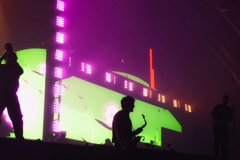 Silhouetted figures against a neon green and purple illuminated industrial backdrop during a nighttime performance.