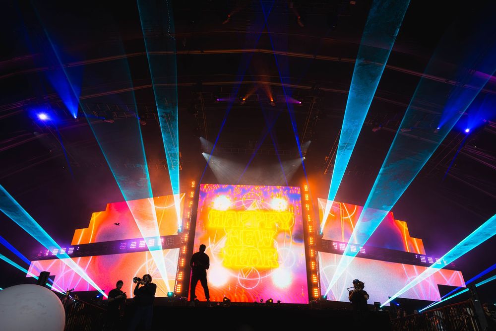 Silhouettes of concert crowd against vibrant blue laser lights and colorful stage production during live performance.