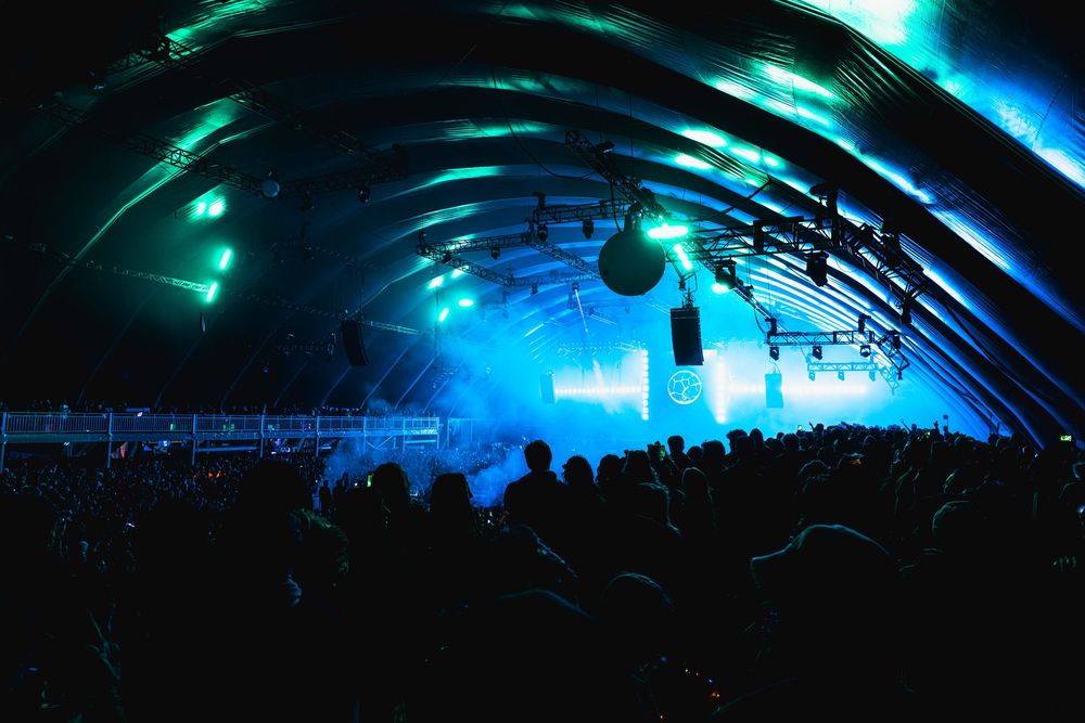 Silhouetted concert crowd under illuminated dome with dramatic turquoise and blue lighting effects at music festival.