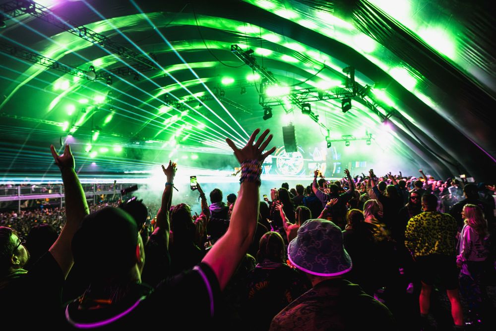 Concert crowd silhouettes with raised arms under green laser lights and illuminated dome ceiling at electronic music festival.