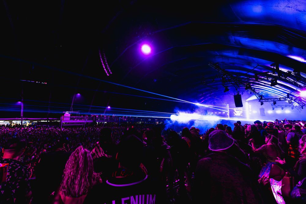 A large crowd at a nighttime concert venue with purple lighting and bright beams illuminating the dark atmosphere.