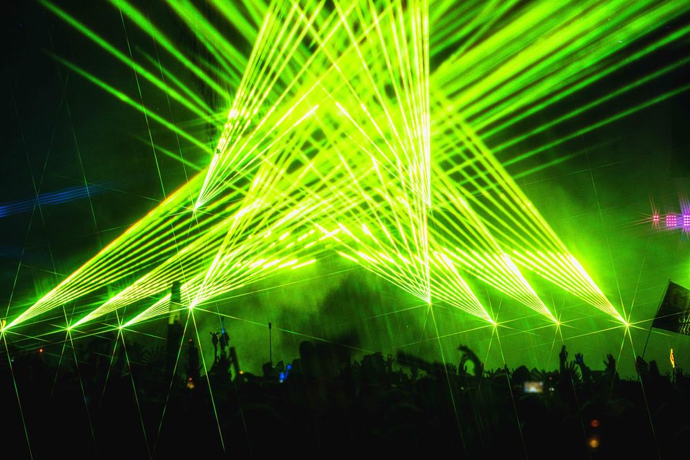 Vibrant green laser beams create geometric patterns across a dark concert venue ceiling during an electronic music performance.