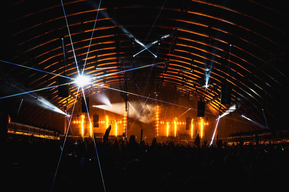 Concert venue illuminated with dramatic orange lighting and laser beams under an arched ceiling during a live performance.