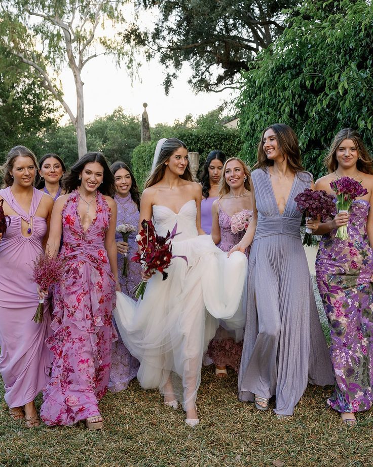 Bride surrounded by bridesmaids in purple and lavender floral gowns outdoors.