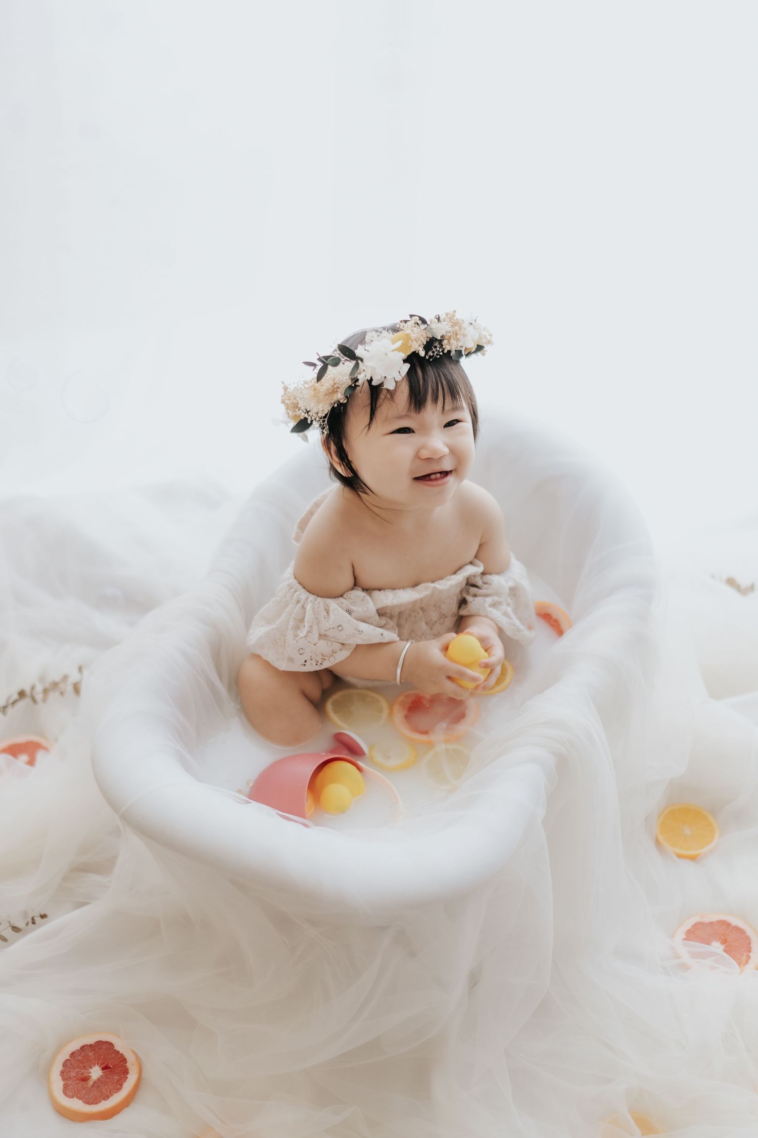 Baby wearing floral crown sits in white tulle dress surrounded by citrus fruit slices for ethereal photo session.