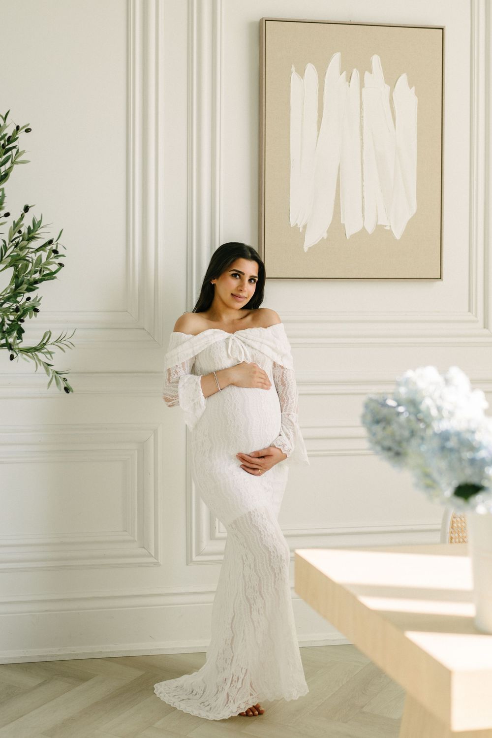 Expectant mother in a fitted lace gown holding white flowers.