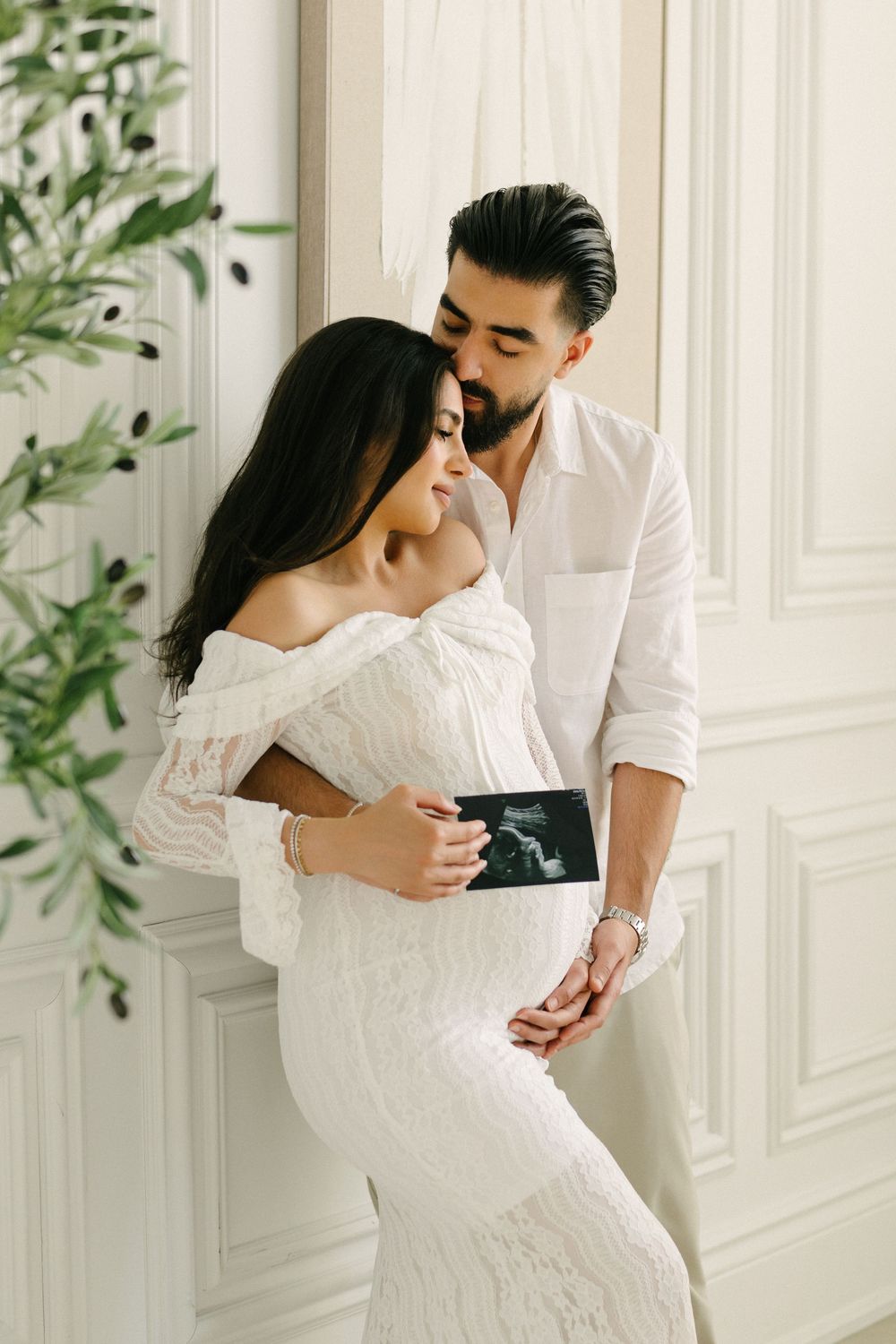 Expectant couple embracing beside a doorway with greenery.