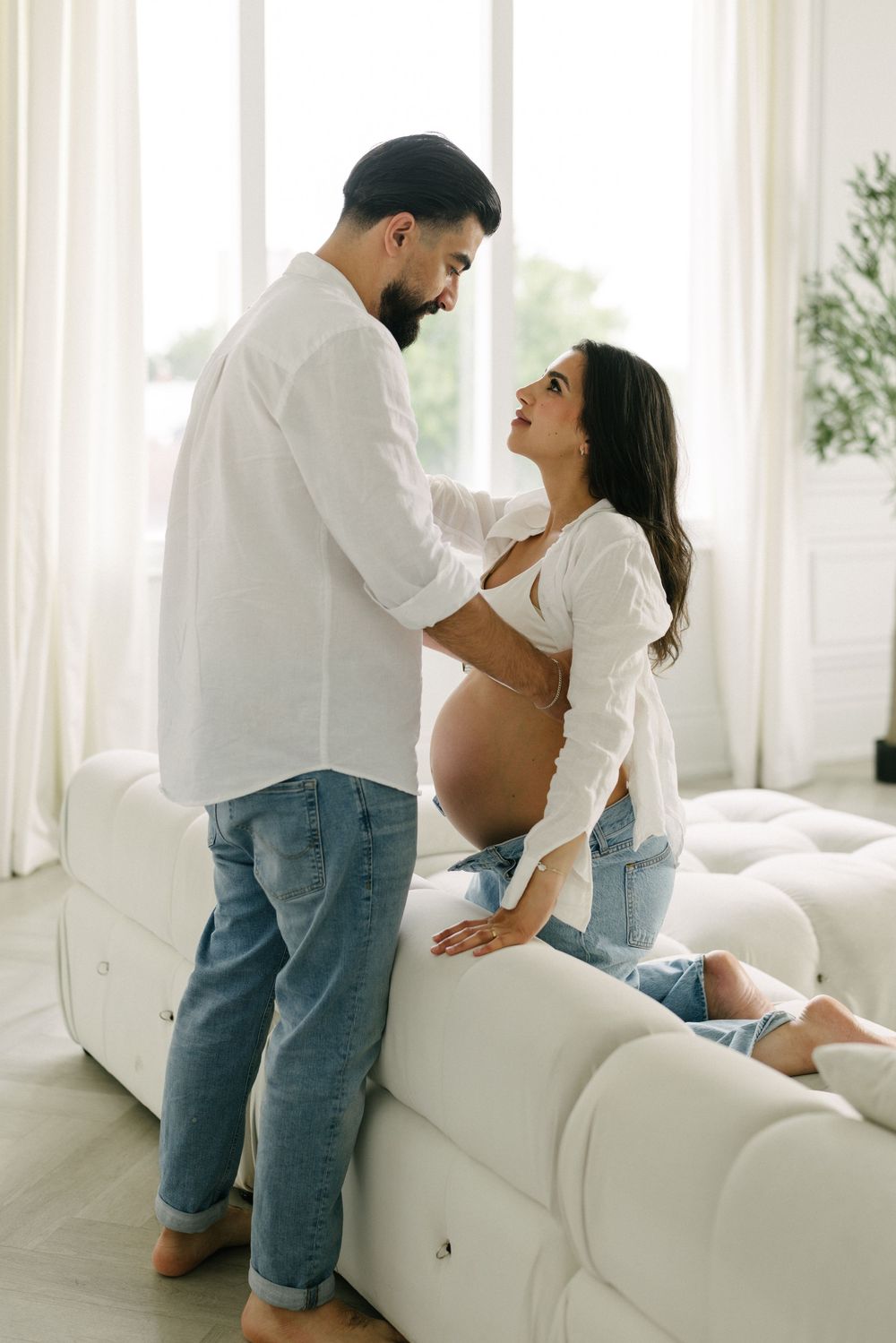 Expectant couple standing together beside a white sofa.