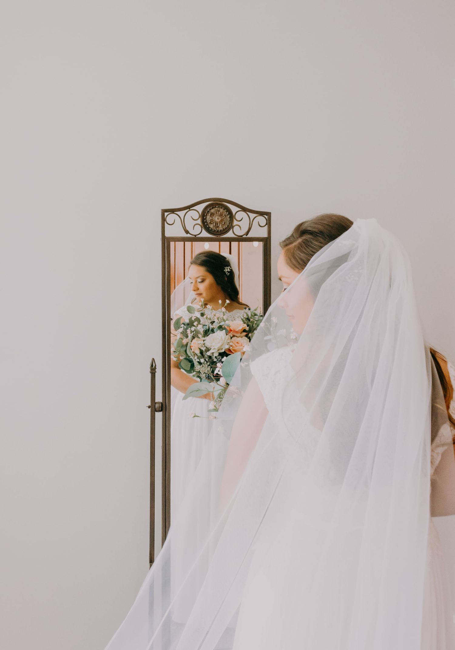 Bride in a white gown and veil holding a bouquet while gazing into an ornate mirror.