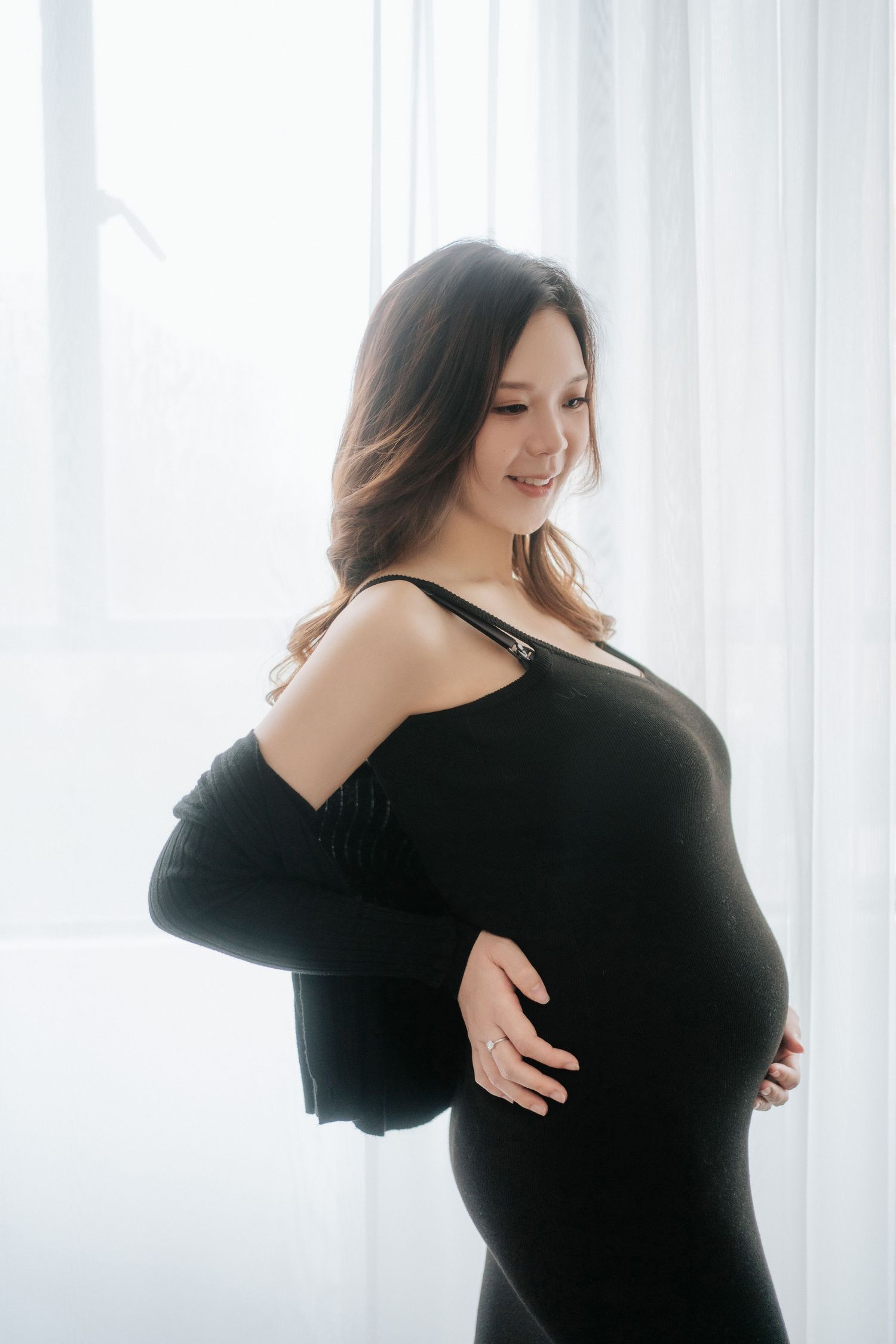 A silhouetted figure in a black dress poses gracefully against bright white curtains in a maternity photoshoot.