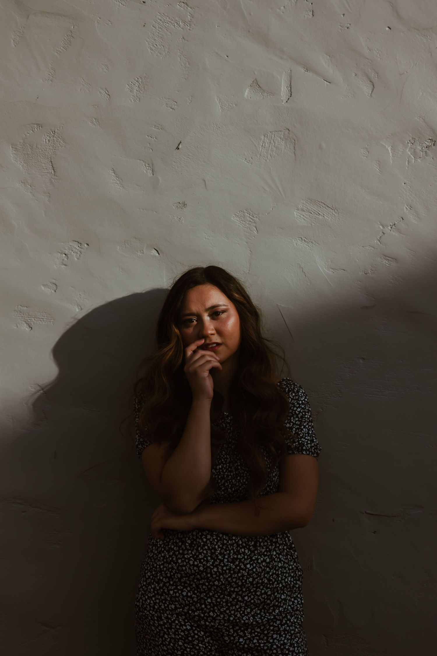 Young woman in floral dress leaning against textured white wall in dramatic moody lighting portrait.