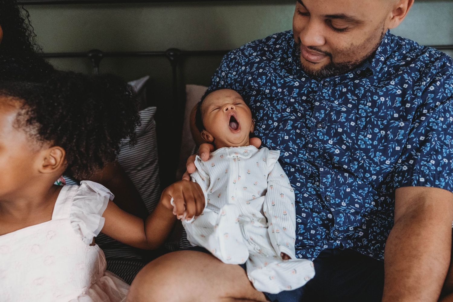 A father holds a yawning newborn baby while a young girl looks on during a warm family moment indoors.