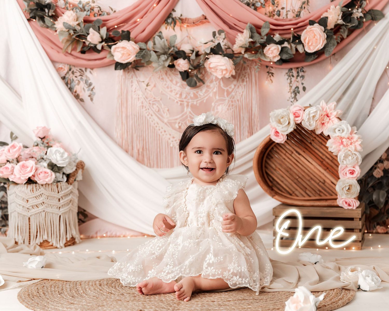 Baby girl in white lace dress sitting at boho floral backdrop for first birthday cake smash photo.