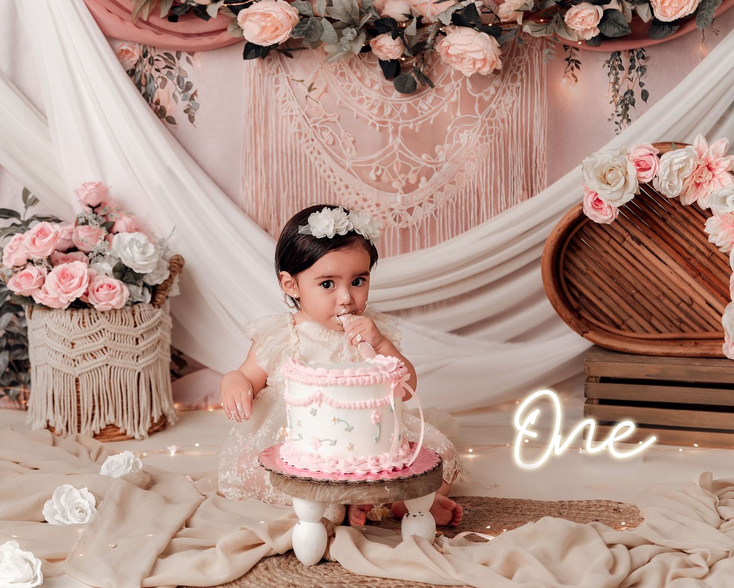 Baby girl in white dress sits before pink floral cake at first birthday smash cake photo session.