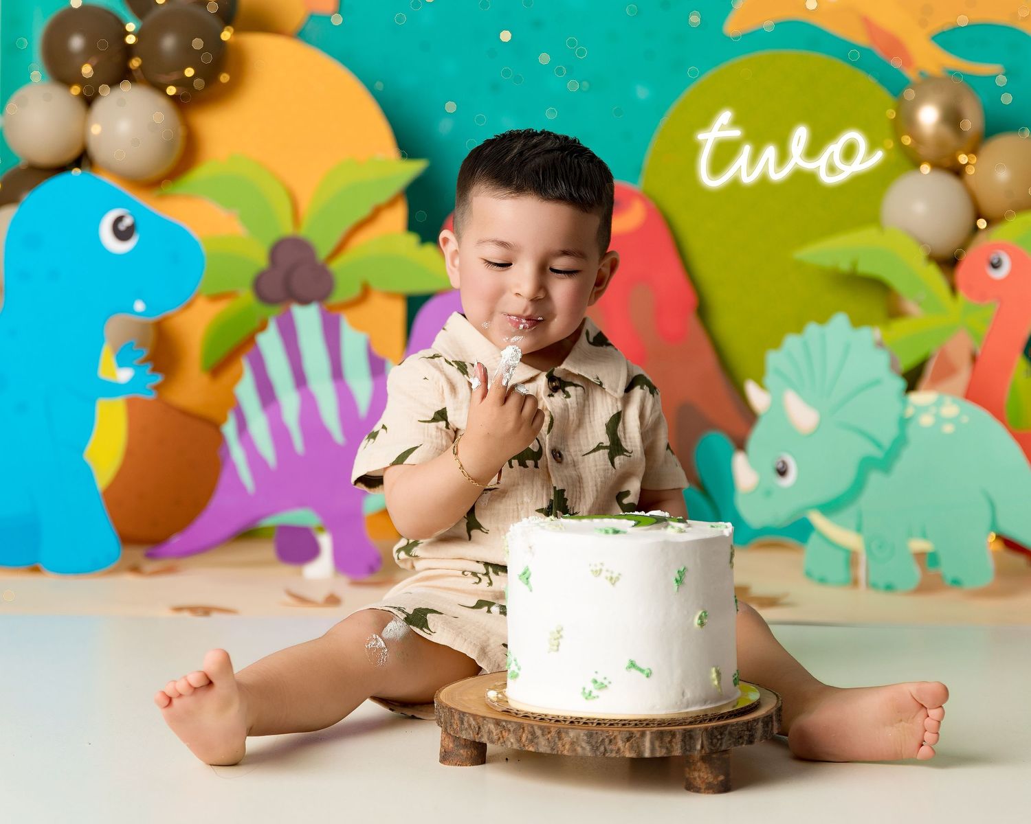 A toddler boy celebrates his second birthday with a dinosaur-themed cake smash, surrounded by colorful dino decorations.