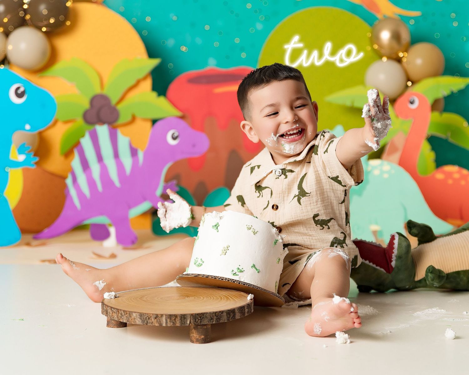 Smiling toddler celebrates second birthday with dinosaur cake smash, surrounded by colorful dino decorations and balloons.