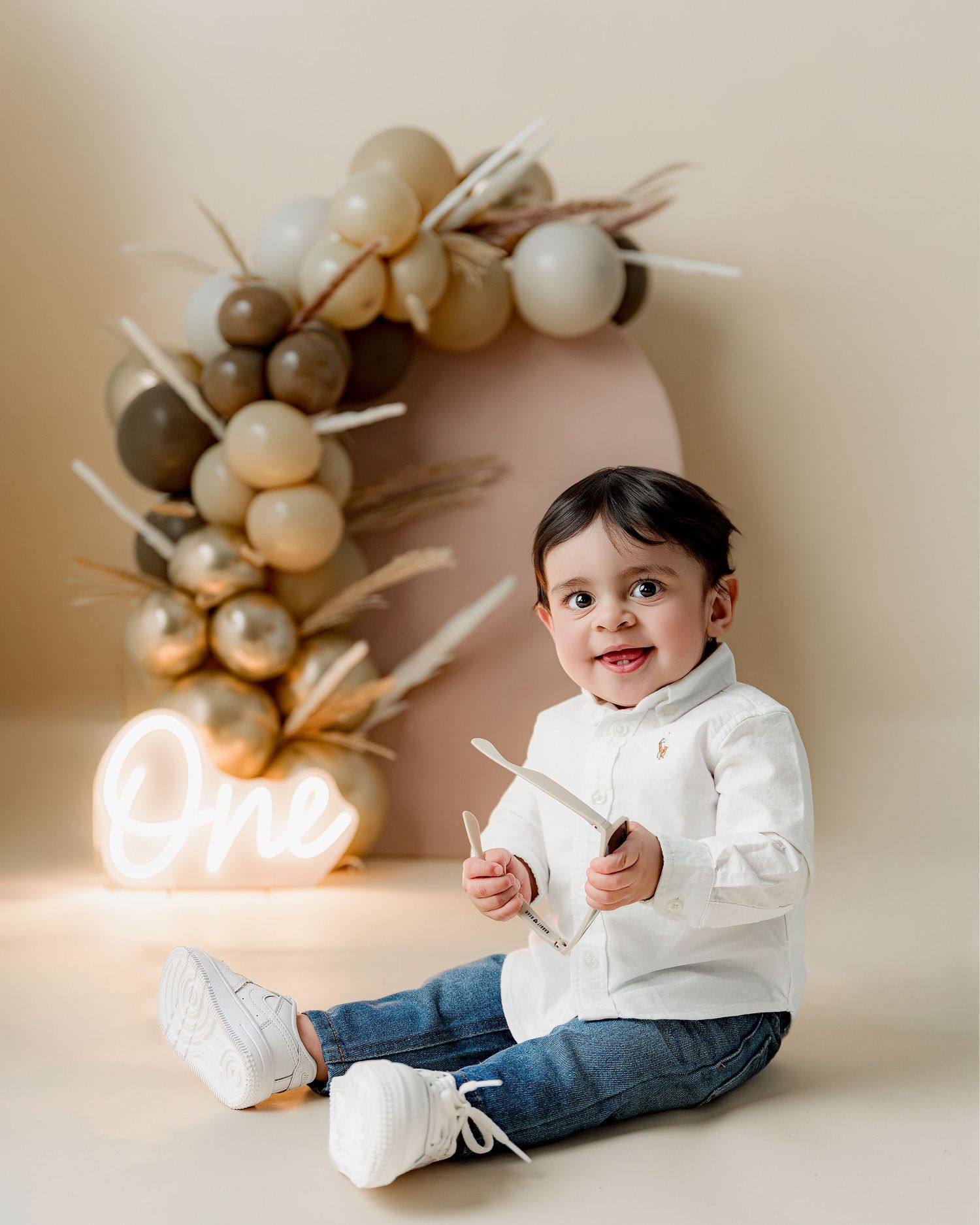 A smiling toddler boy in white shirt and jeans sits before a balloon arch with a glowing One neon sign.