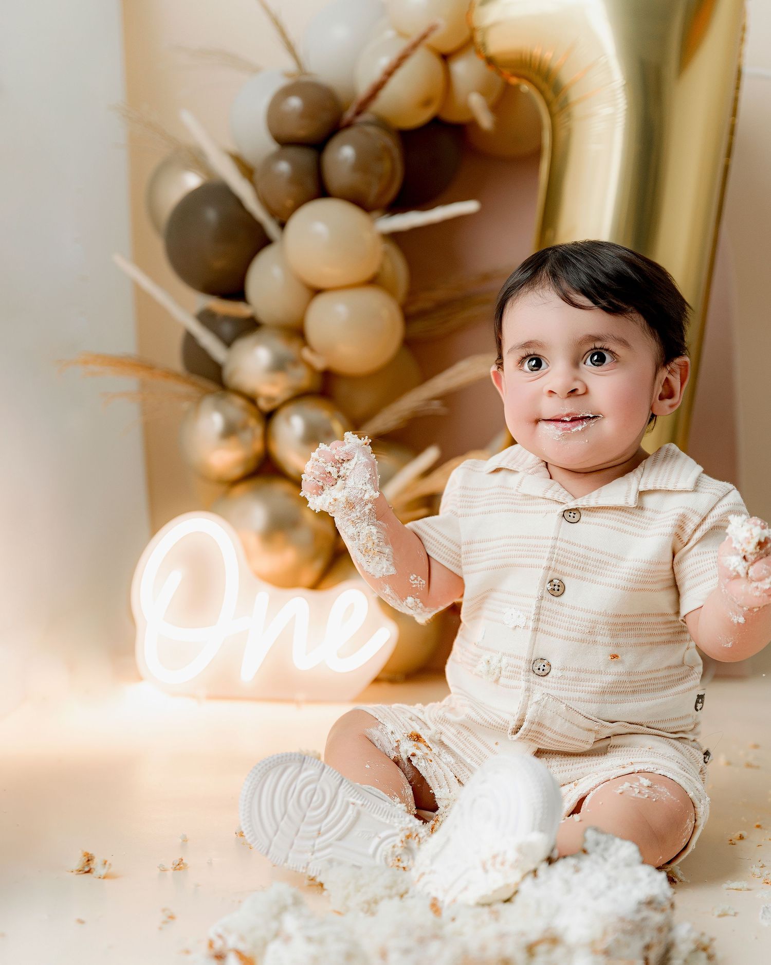 Baby boy in beige outfit celebrates first birthday smash cake with balloon backdrop and neon One sign.