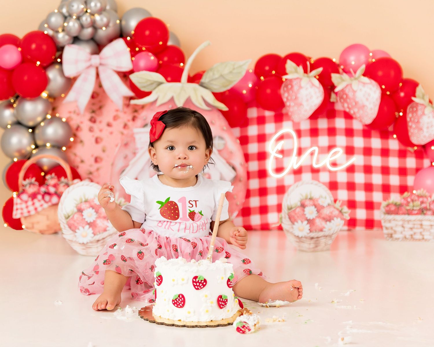 Baby girl in strawberry outfit celebrates first birthday with a smash cake at a festive strawberry-themed photo shoot.