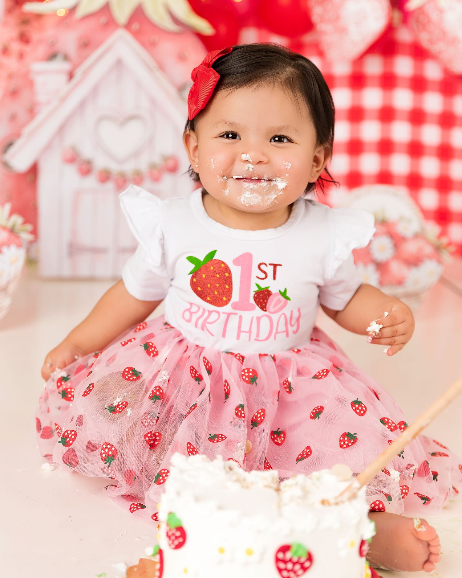 Baby girl in strawberry 1st birthday outfit smashes cake, wearing pink tutu dress with frosting on her face.