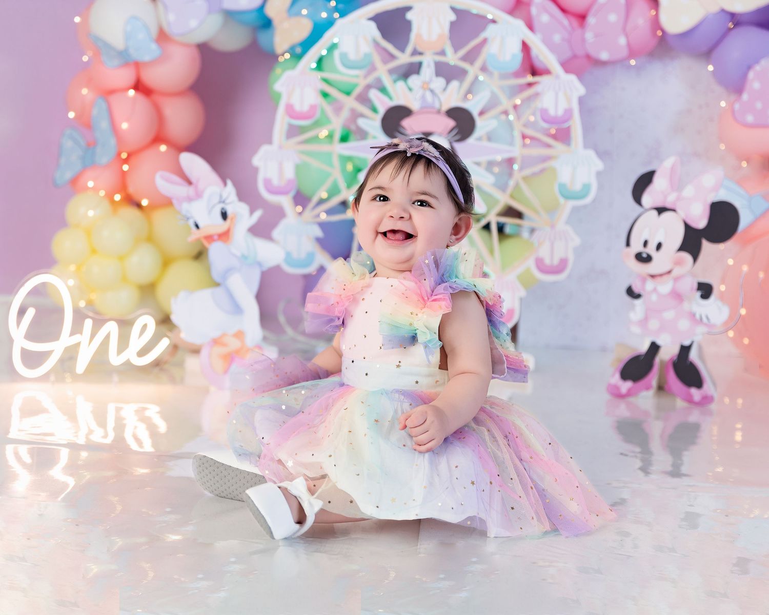 A baby girl in a rainbow tutu dress celebrates her first birthday with Minnie Mouse balloons and decorations.