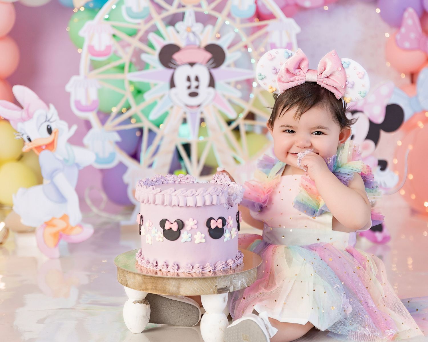 Baby girl in rainbow tutu celebrates 1st birthday with Minnie Mouse themed cake smash and colorful balloon backdrop.