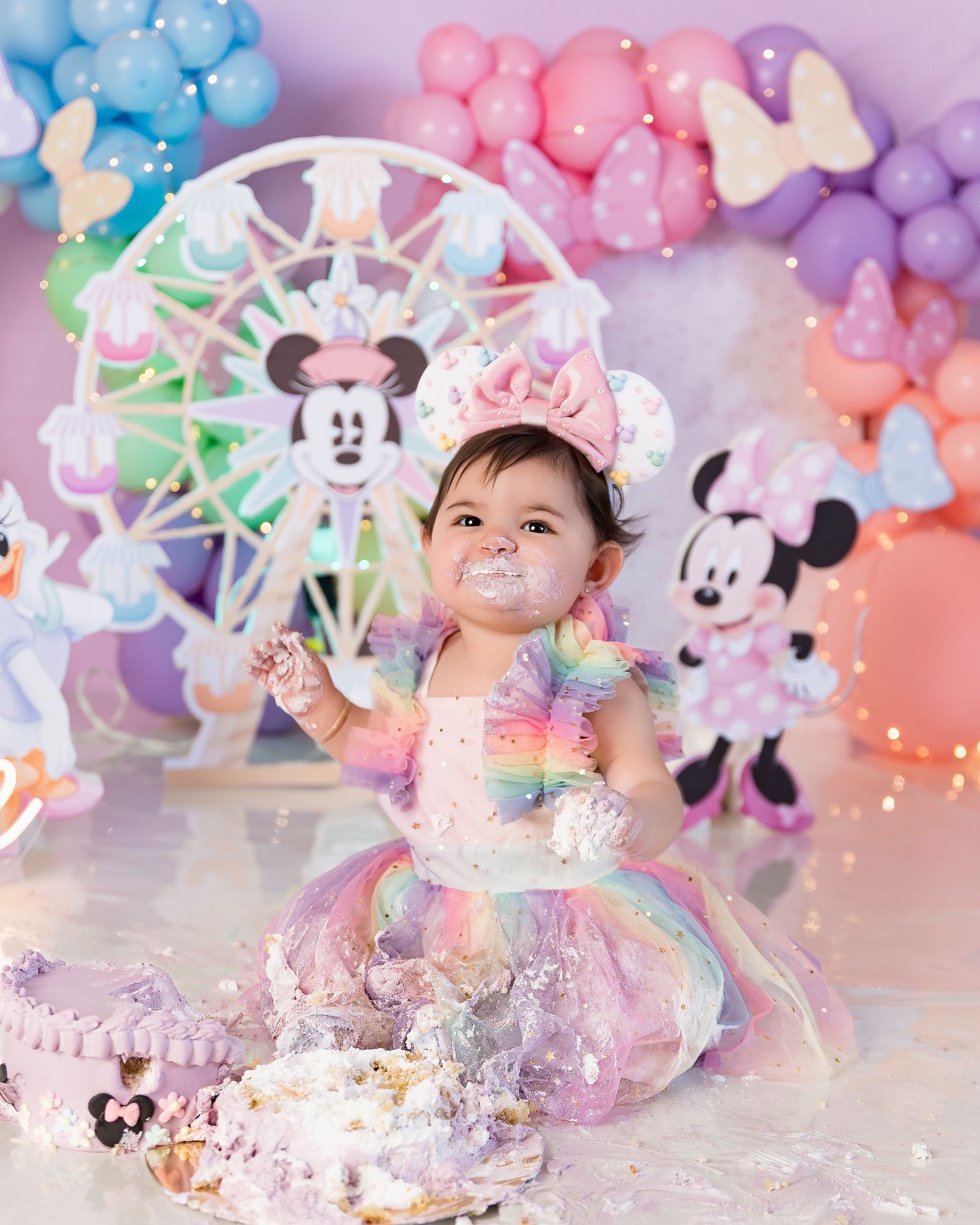 Baby girl in rainbow tutu smashes first birthday cake at Minnie Mouse themed party with colorful balloon arch.