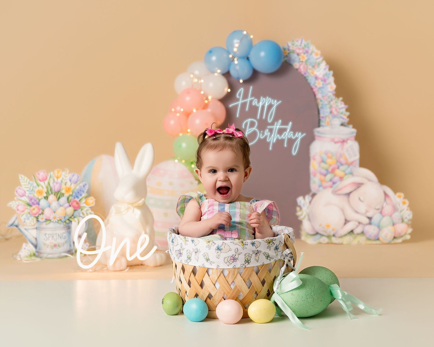Baby girl in basket with Easter eggs, bunnies, and Happy Birthday sign for a first birthday photo.