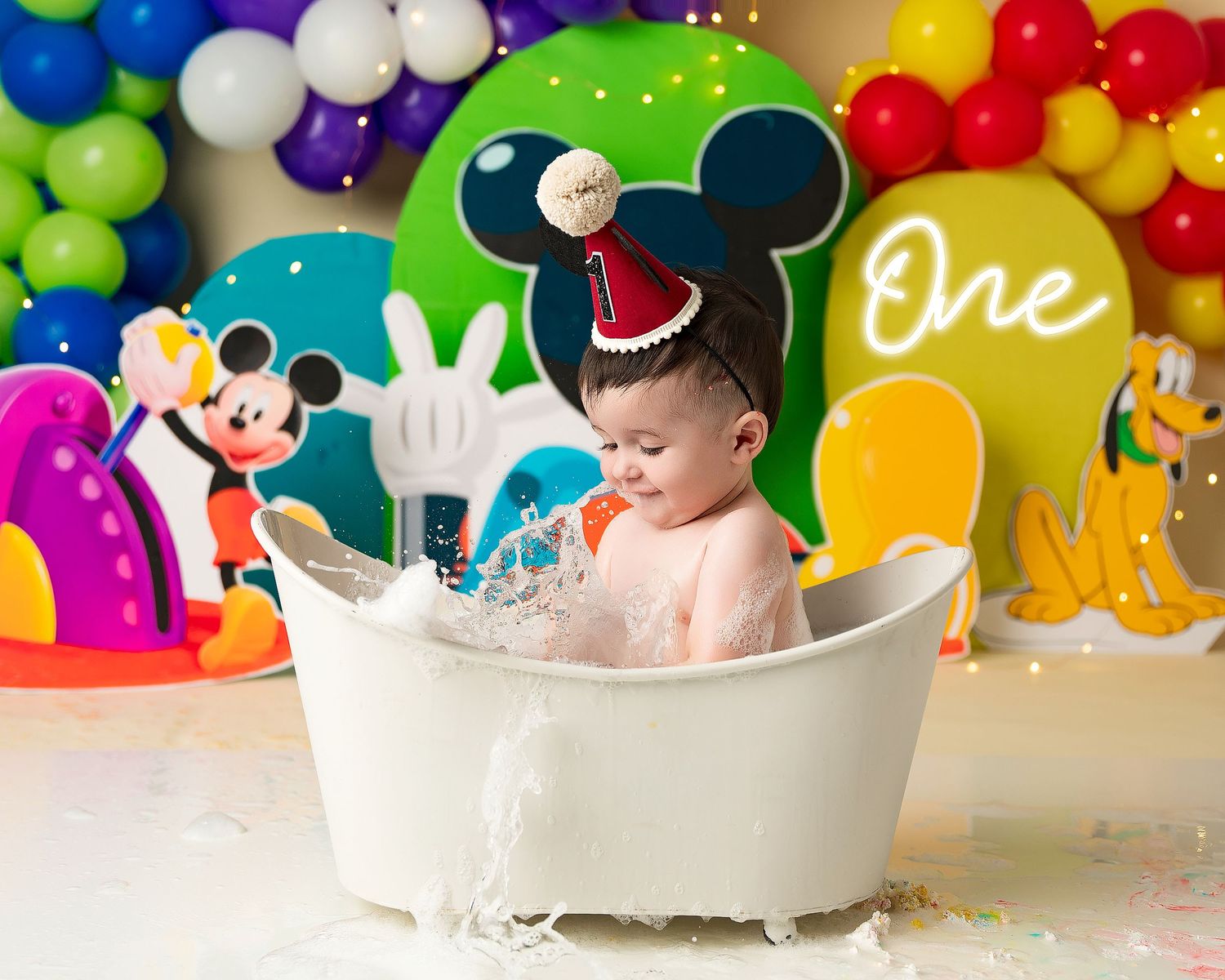 A colorful first birthday cake smash photo session featuring a white bathtub and rainbow balloon backdrop.