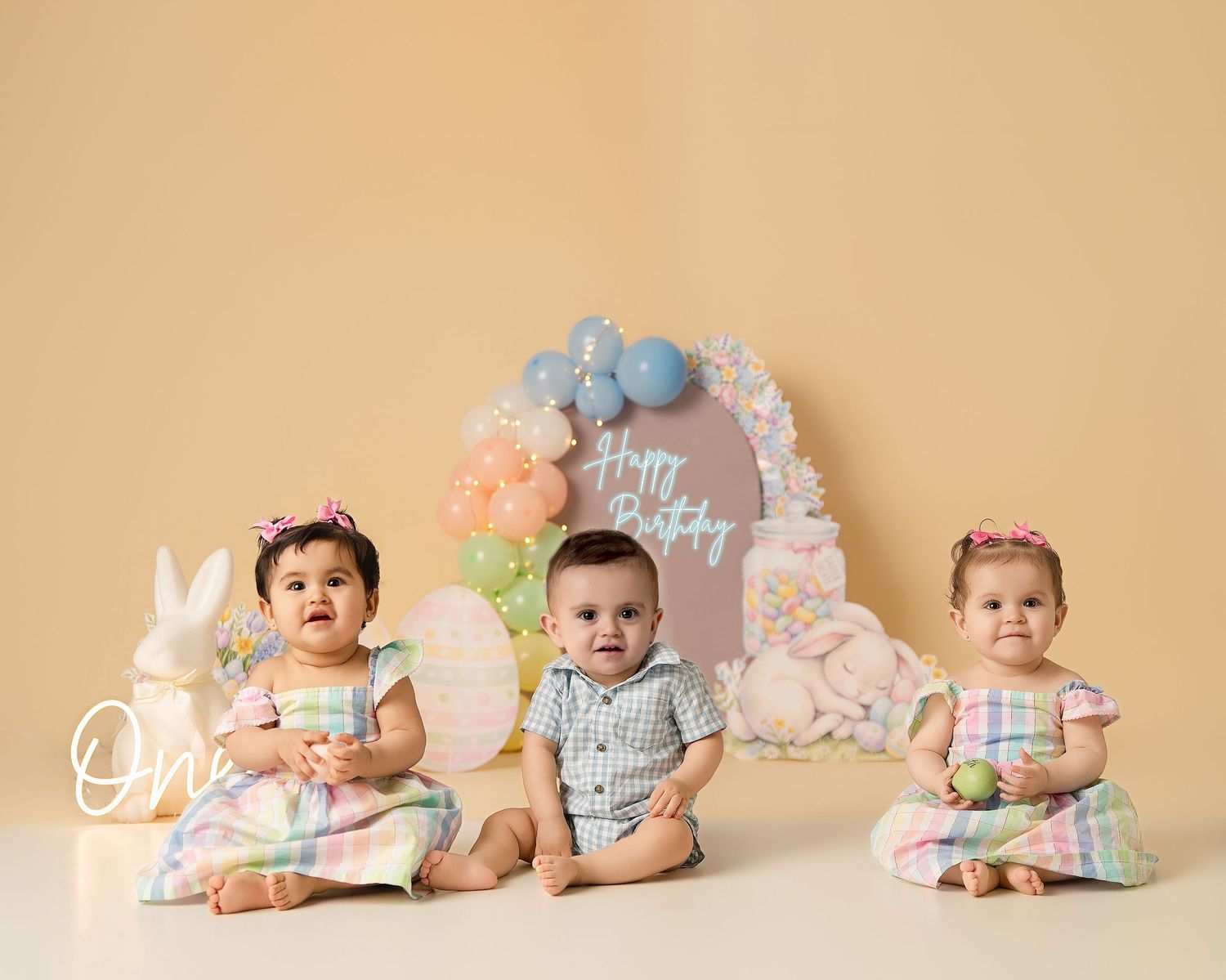 Three babies sit together in front of a Happy Birthday arch with balloons and a white bunny decoration on a beige backdrop.