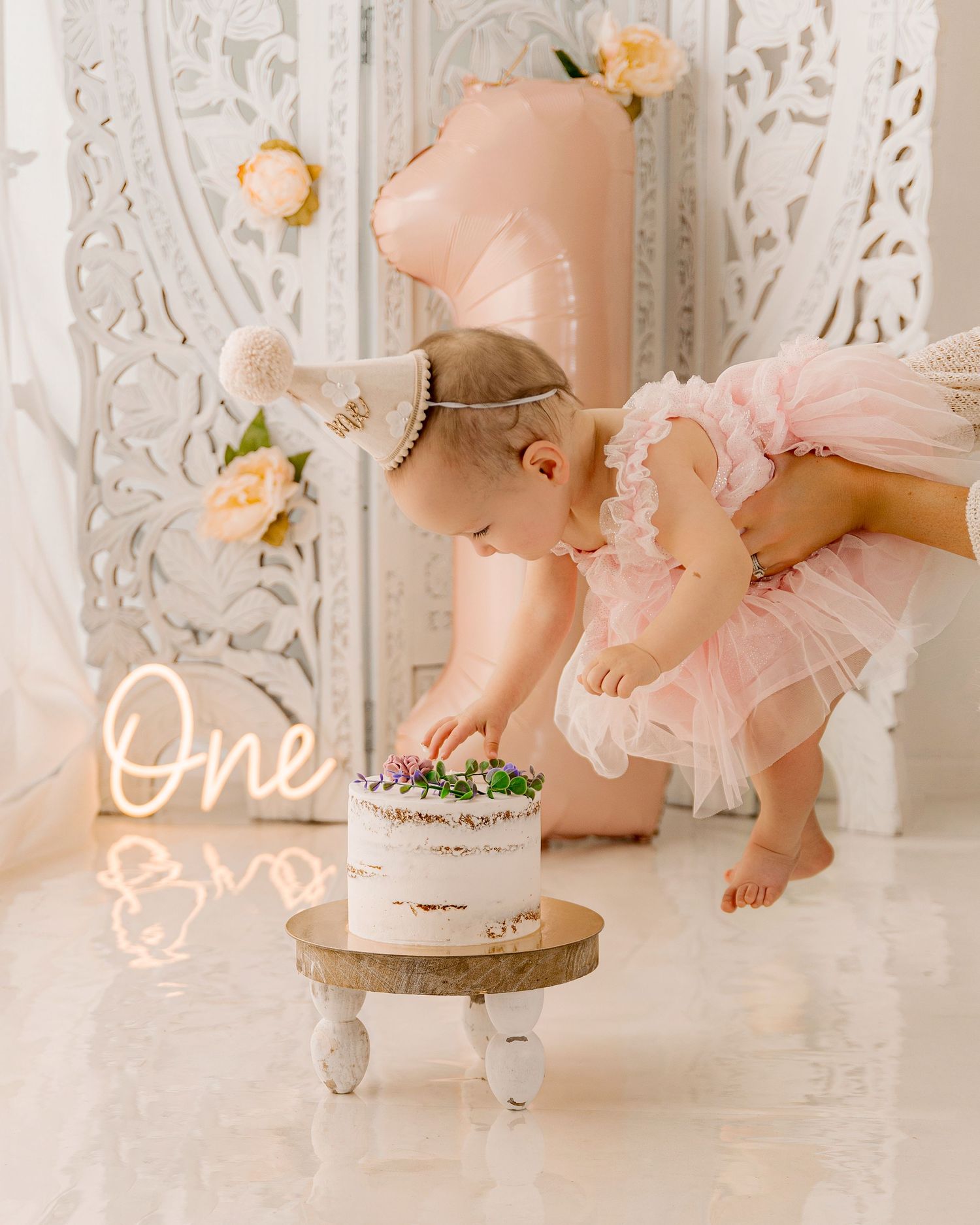 Baby girl in pink tutu smashes first birthday cake beside a glowing "One" neon sign and number 1 balloon.