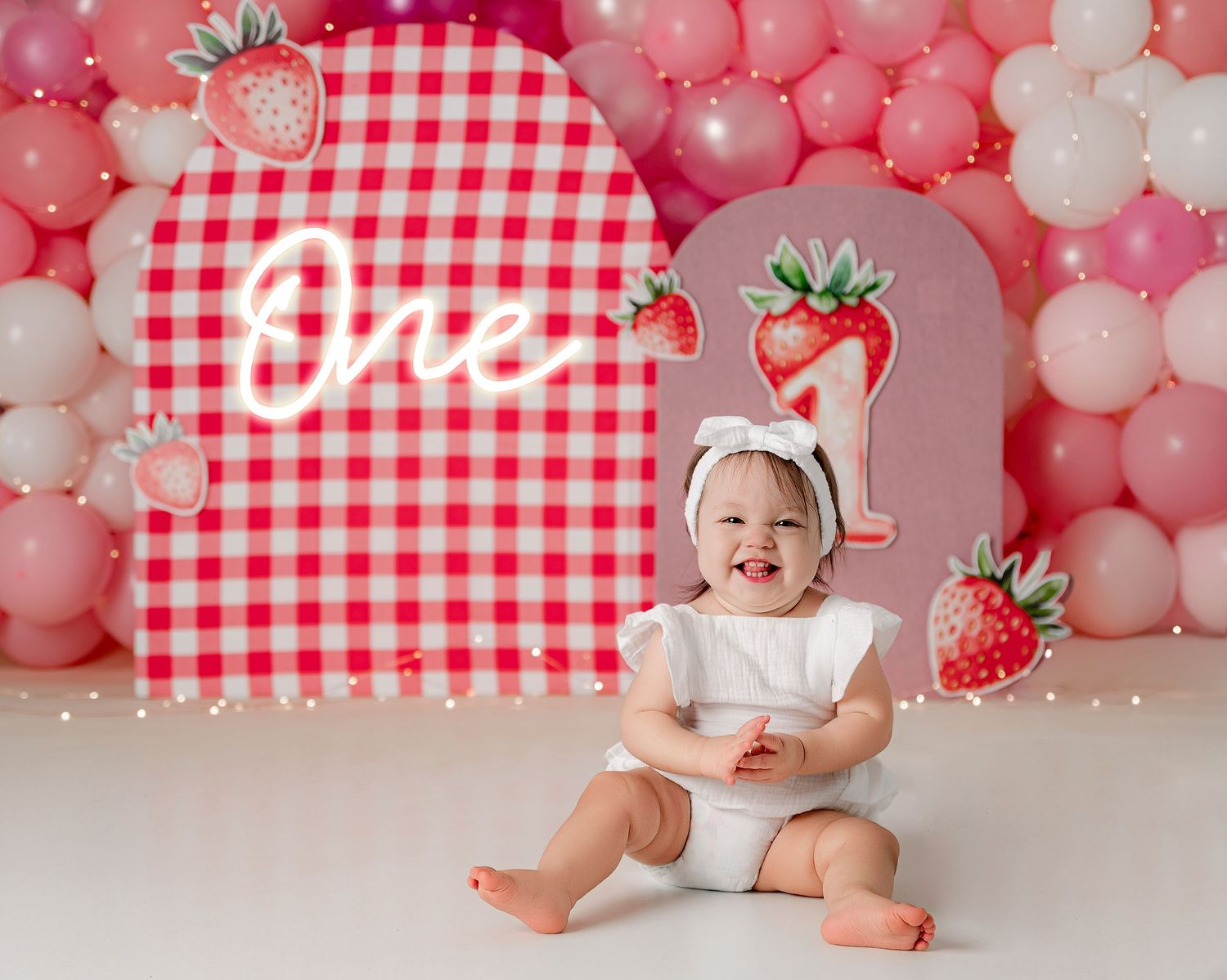 Red and white gingham backdrop with pink and white balloon arch creates a sweet strawberry-themed first birthday setting.