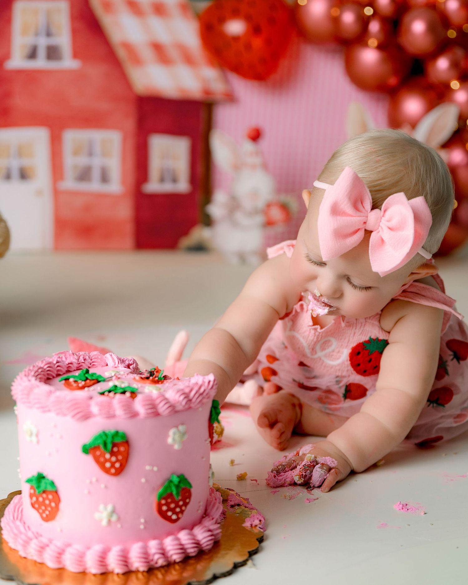 A pink frosted birthday cake with strawberry decorations sits on a table while someone smashes their face into it.
