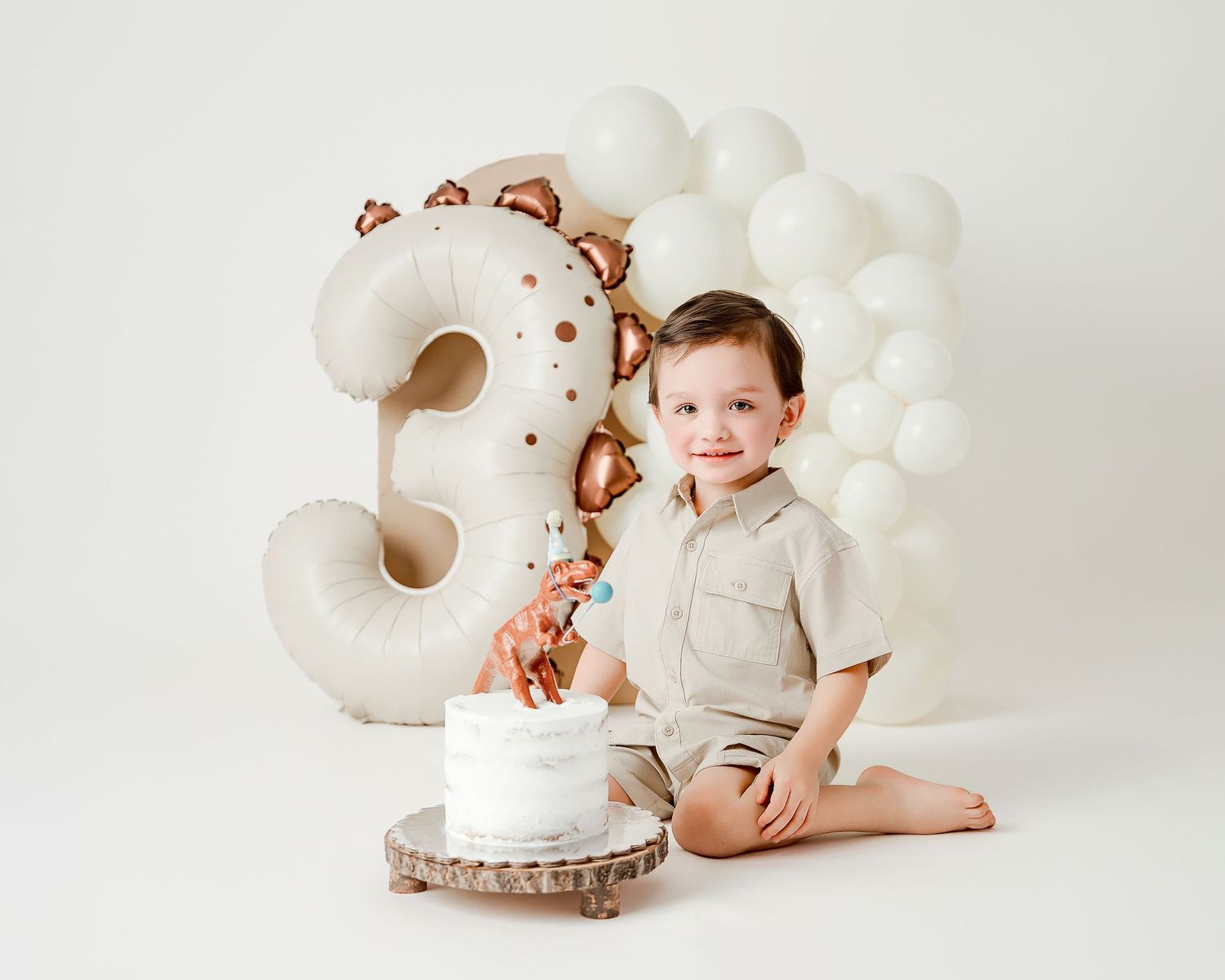 Toddler boy sitting with a white cake in front of a number 3 balloon and white balloon arch for a third birthday.