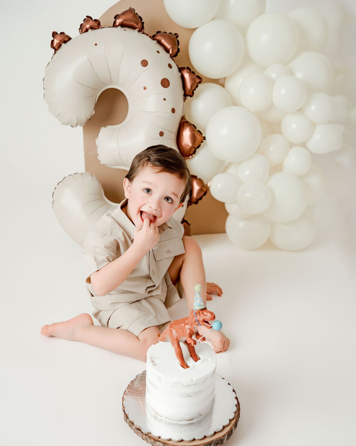 Toddler boy smashing white dinosaur cake at a 2nd birthday party with white balloons and number 2 balloon backdrop.