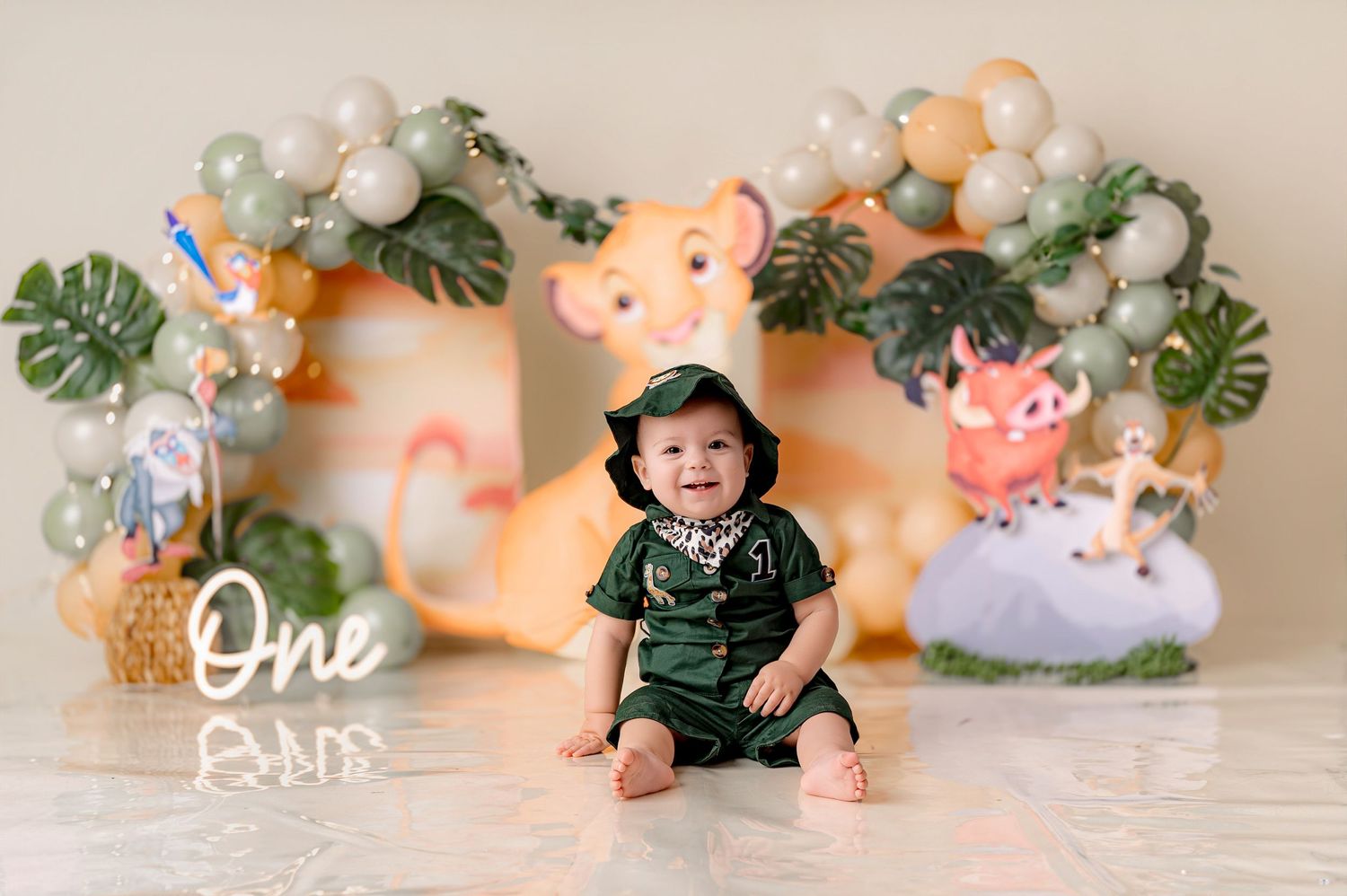 Baby's first birthday photoshoot with Lion King theme, balloons, and tropical decor, smiling infant in green outfit.