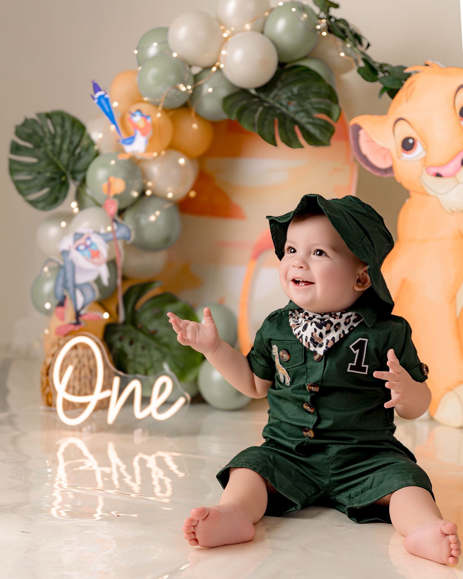 Baby boy in green outfit celebrates first birthday with Lion King themed decorations and balloon arch.